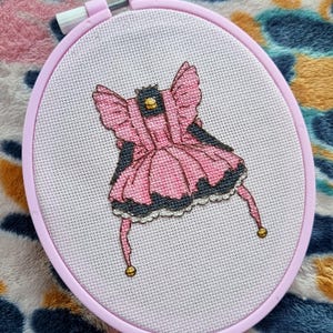 May include: A pink and black embroidered dress with gold accents, framed in a pink embroidery hoop.