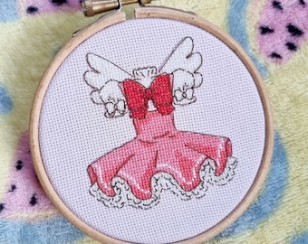 Op de afbeelding: Een roze en witte kruissteekborduursel van een jurk met vleugels en een rode strik. De borduursel is ingelijst in een houten hoepel.