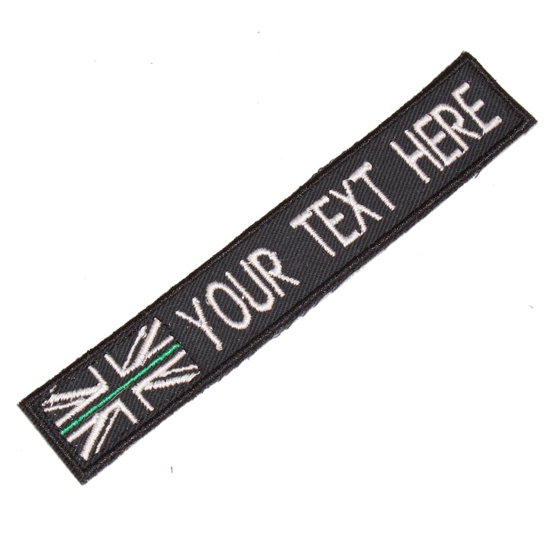 May include: Black embroidered patch with a white Union Jack flag with a green stripe across the middle and the text "YOUR TEXT HERE" in white.