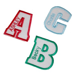 May include: Three felt letters, A, B, and C, with white fabric and coloured embroidered edges. The letter A is red, the letter B is green, and the letter C is blue. Each letter has a name embroidered on it: Ashley, Becky, and Charlie.