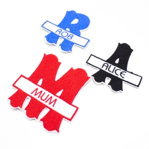 May include: Three embroidered iron-on patches with names in white lettering on a white background. The patches are shaped like letters, with the names "Rob", "Alice", and "Mum" on them. The letters are blue, black, and red, respectively.