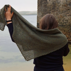 May include: A person wearing a green crocheted shawl with a scalloped edge. The shawl is draped over the person's shoulders and is held up by one hand. The person is standing in front of a stone wall and a body of water.