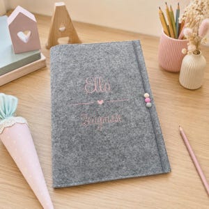 Könnte beinhalten: Eine graue Filzmappe mit dem eingestickten Namen Ella und dem Wort Zeugnisse in rosa Garn, getrennt durch ein kleines rosa Herz. Die Mappe wird mit einem Gummiband verschlossen, das mit drei kleinen, runden, bunten Filzkugeln verziert ist.