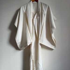 May include: Off-white kimono-style robe hanging on a wooden hanger. The robe has wide sleeves and a draped front. The fabric appears to be a lightweight material, possibly silk or a similar fabric. The robe is hanging against a plain white wall.