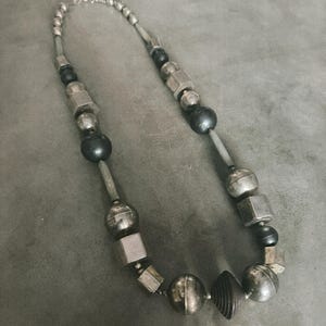 Puede incluir: Un collar que presenta una secuencia de cuentas metálicas plateadas alternadas con cuentas esféricas de color gris oscuro. El diseño incorpora varias formas geométricas, incluyendo cubos, esferas y una cuenta cónica estriada central.