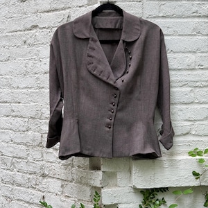 Könnte beinhalten: Eine dunkelbraune Jacke mit langen Ärmeln und abgerundetem Kragen, die einen diagonalen Knopfverschluss auf der Vorderseite sowie Ziernähte am Revers aufweist. Die Jacke hat eine taillierte Passform, einen leicht ausgestellten Saum und Ärmelaufschläge.