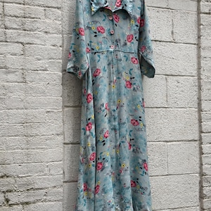 May include: A long-sleeved, floor-length dress in a light blue hue, featuring a repeating floral pattern with pink roses, yellow blossoms, and green leaves. The garment includes a pointed collar, a button-down front closure, and a flowing skirt.