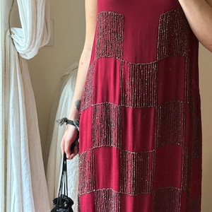 May include: A sleeveless, knee-length maroon dress featuring a checkerboard pattern created with dark, metallic beadwork. The garment has a round neckline accented with matching beadwork and a straight, relaxed silhouette throughout the body.