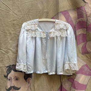 Könnte beinhalten: Eine hellblaue Jacke mit langen Ärmeln und V-Ausschnitt, verziert mit cremefarbener Spitzenborte an den Schultern und Ärmelbündchen. Das Kleidungsstück hat ein schmales Stoffband als Verschluss in der vorderen Mitte und hängt an einem weißen Kleiderbügel.