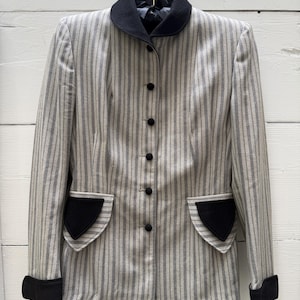 Puede incluir: Una chaqueta de manga larga con un patrón de rayas verticales en gris claro y gris oscuro. La prenda incluye un cuello redondeado en negro sólido, botones negros, puños negros y dos solapas de bolsillo decorativas en color negro al frente.