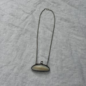 May include: A silver-toned metal necklace featuring a thin chain and a pendant shaped like a small, elongated, curved purse or coin holder. The pendant has a smooth, cream-coloured, shell-like inlay with a dark metal frame and a small clasp at the top.