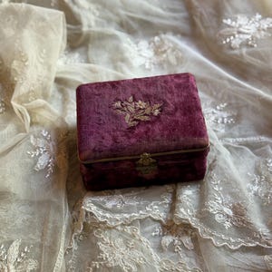 May include: A small, rectangular, burgundy velvet box with a gold-colored floral embroidery on the lid. The box has a gold-colored clasp and hinges. The box is set on a background of delicate, cream-colored lace.