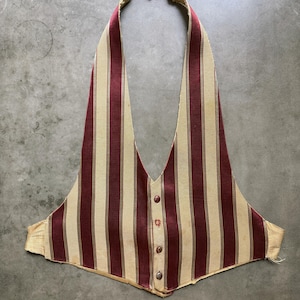 Puede incluir: Un chaleco sin mangas con un patrón de rayas verticales en rojo oscuro y crema. La prenda tiene un escote en V profundo, una tira para el cuello estilo halter y un cierre frontal con cuatro botones circulares pequeños. El dobladillo termina en punta.