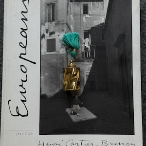 May include: A gold-toned metal object with a twisted cylindrical body and a domed lid, topped with a bright teal tassel. The object is placed on the cover of a book titled Europeans by Henri Cartier-Bresson, which features a black and white photograph.