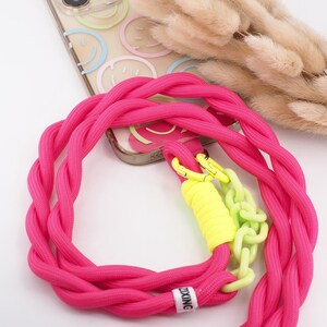 Könnte beinhalten: Ein leuchtend pinkfarbenes Telefonband mit geflochtenem Seildesign. Das Band hat gelbe und grüne Akzente, darunter eine Kette und ein zylindrisches Element. Eine transparente Handyhülle mit Smiley-Gesichtern ist ebenfalls sichtbar.