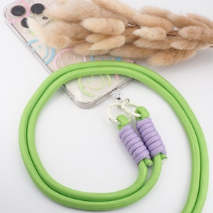 Könnte beinhalten: Ein grünes Handyband mit einem lila geflochtenen Band und silbernen Verschlüssen. Das Band ist an einer durchsichtigen Handyhülle mit einem bunten Smiley-Design befestigt.