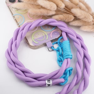 Könnte beinhalten: Ein lila geflochtenes Handyband mit einer blauen Kette und einer durchsichtigen Handyhülle mit einem Smiley-Design. Das Band hat ein Etikett mit der Aufschrift "NZK".