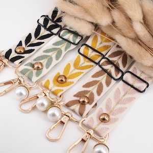 Könnte beinhalten: Fünf dekorative Schlüsselanhänger mit gestickten Blattmustern in Schwarz, Grün, Gelb, Braun und Rosa auf cremefarbenem Hintergrund. Jeder Schlüsselanhänger hat einen goldenen Verschluss und einen Perlenakzent. Die Schlüsselanhänger sind in einer Reihe angeordnet.