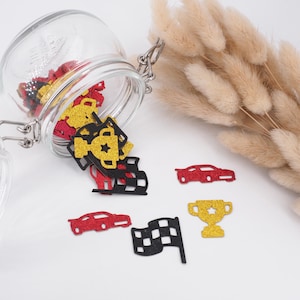 Könnte beinhalten: Ein Glasgefäß gefüllt mit rotem, schwarzem und goldenem Glitzerkonfetti. Das Konfetti ist in Form von Rennwagen, karierten Flaggen und Trophäen. Das Gefäß verschüttet das Konfetti auf eine weiße Oberfläche.