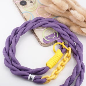 Könnte beinhalten: Eine transparente Handyhülle mit Smiley-Design wird mit einem geflochtenen lila Handyband kombiniert. Das Band hat eine gelbe Kette und ein gelbes Seildetail mit einem Metallverschluss. Die Handyhülle befindet sich auf einer weißen Oberfläche.