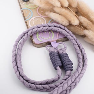 Könnte beinhalten: Ein geflochtener, lavendelfarbener Handyriemen mit grauen Akzenten und eine transparente Handyhülle mit Smiley-Gesichtern in Pastellfarben. Der Riemen ist mit einem kleinen Metallverschluss an der Handyhülle befestigt.