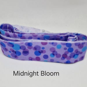 May include: A fabric item with a lavender background and a pattern of blue and purple circles. The text "Midnight Bloom" is printed at the bottom of the image. The item is folded over on itself.