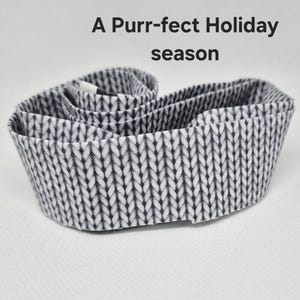 May include: A coiled fabric strip featuring a black and white printed pattern that mimics the appearance of a knitted texture. Above the fabric, the text reads A Purr-fect Holiday season in a simple, dark gray sans-serif font.