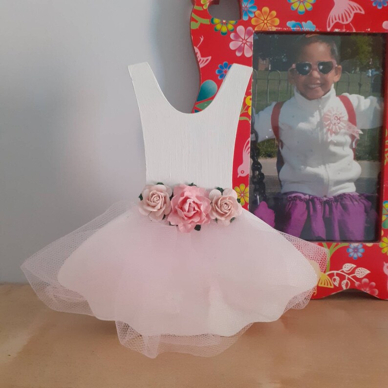 May include: A white wooden ballerina dress cutout with a pink tulle skirt and three pink paper roses. A red floral picture frame with a photo of a child is in the background.