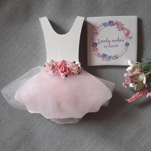 May include: A white wooden ballerina dress cutout with a pink tulle skirt and pink paper roses. A small square tile with the text "Lovely makes by Rachel" and a floral design is next to the dress.