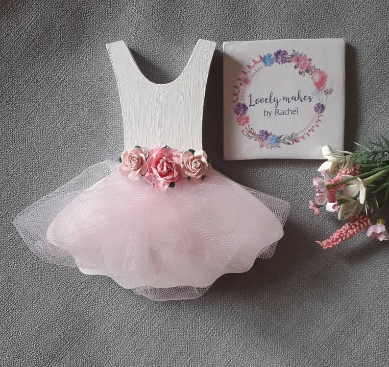 May include: A white wooden ballerina dress cutout with a pink tulle skirt and pink paper roses. A small square tile with the text "Lovely makes by Rachel" and a floral design is next to the dress.