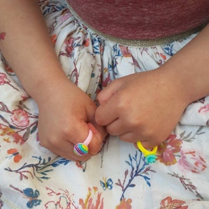 May include: A child's hands wearing two colourful plastic rings. The rings are decorated with stripes and a flower design. The child is wearing a white dress with a floral pattern.
