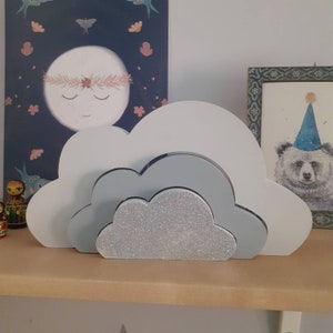 Puede incluir: Una estantería decorativa con forma de nube en tonos blanco, gris y purpurina plateada. El diseño en capas presenta una nube brillante en el centro, con nubes más grandes en blanco y gris. La estantería está sobre una superficie de madera.