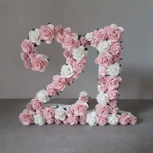 Puede incluir: Un número decorativo "21" hecho de material blanco, adornado con rosas de papel rosas y blancas. El arreglo floral crea un efecto tridimensional, ideal para celebraciones. El número mide aproximadamente 30 cm de alto.