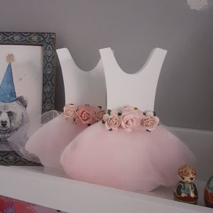 May include: Two white wooden dress-shaped decorations with pink tulle skirts and floral waistbands. The dresses are decorated with pink roses and are displayed on a white shelf. A framed bear illustration is visible in the background.