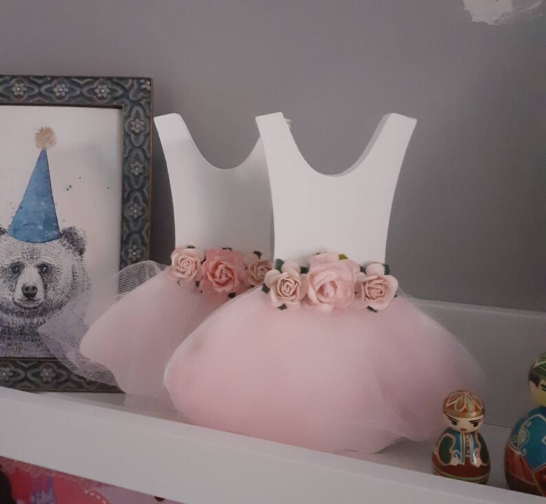 May include: Two white wooden dress-shaped decorations with pink tulle skirts and floral waistbands. The dresses are decorated with pink roses and are displayed on a white shelf. A framed bear illustration is visible in the background.