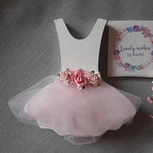 May include: A wooden ballerina dress cutout painted white, with a pink tulle skirt and pink paper roses at the waist. The text "Lovely makes by Rachel" is on a small square block.
