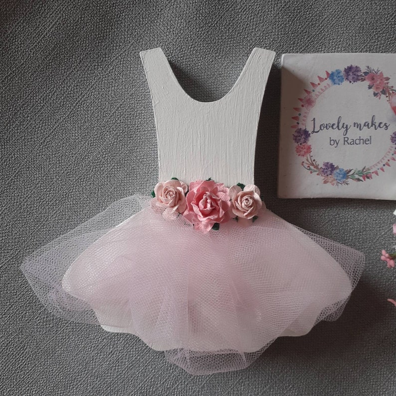 May include: A wooden ballerina dress cutout painted white, with a pink tulle skirt and pink paper roses at the waist. The text "Lovely makes by Rachel" is on a small square block.