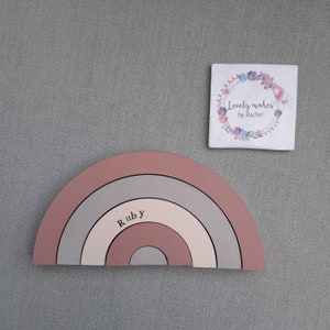 May include: A wooden rainbow in shades of pink, gray, and cream, with the name "Ruby" printed on it. A small square sign with floral wreath and text "Lovely makes by Rachel" is also present.