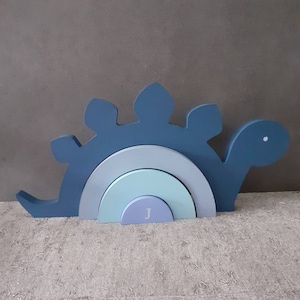 May include: A wooden dinosaur stacking toy in shades of blue, grey, and mint green. The toy features a curved rainbow design and a white initial "J" on the bottom piece. The dinosaur has a friendly face and is designed for children.