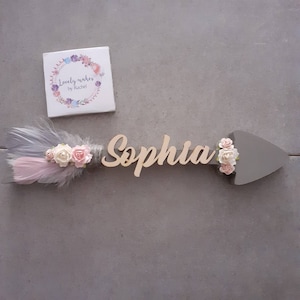 Puede incluir: Una flecha de madera con el nombre "Sophia" escrito en ella. La flecha está decorada con flores rosas y blancas y una pluma gris.