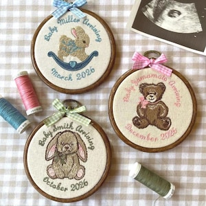 May include: Three circular wooden embroidery hoops featuring fabric panels with stitched designs. One shows a bunny on a rocker with text Baby Miller Arriving March 2026, another a teddy bear with Baby Yamamoto Arriving December 2026, and one a rabbit with Baby Smith Arriving October 2026.