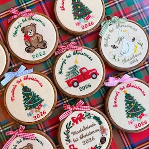 May include: A collection of circular wooden embroidery hoops featuring fabric centers with hand-stitched designs including a teddy bear, a red truck, a goose, and Christmas trees, each personalized with names and the years 2024, 2025, or 2026.