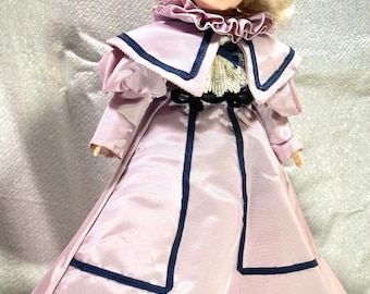 Puede incluir: Una muñeca de porcelana vintage con cabello rubio, vestida con un vestido rosa con ribetes azules y un cuello de encaje blanco.