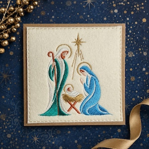 May include: A square greeting card featuring an embroidered nativity scene on a cream-coloured felt patch. The design depicts figures in blue and teal robes surrounding a baby in a manger, with a gold star above, all stitched with detailed thread.