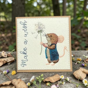 Könnte beinhalten: Gestickte Karte mit einer Maus in einer blauen Weste, die einen Löwenzahn hält. Der Text "Make a wish" steht in Schreibschrift. Die Karte liegt auf einer Holzoberfläche mit Blumen und Blättern.