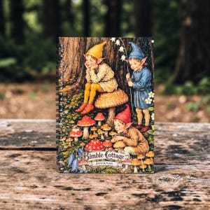 Puede incluir: Un diario con espiral con una ilustración fantástica de gnomos en un bosque. La portada muestra gnomos con gorros puntiagudos, rodeados de setas y el texto "Bimble Cottage Journal."