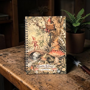 Puede incluir: Un diario con espiral con una ilustración fantástica de gnomos en un bosque. La portada presenta gnomos en un puente, leyendo y cerca de una seta. El diario está etiquetado como "Bimble Cottage Journal".