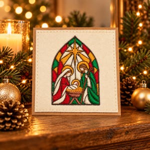 Peut inclure: Une carte de vœux carrée présentant une scène de nativité brodée dans un style vitrail. Le motif montre des personnages en robes rouges et vertes entourant une crèche sous une étoile, le tout encadré par une arche sur un fond en tissu crème.