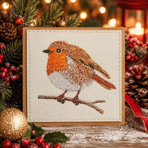 Peut inclure: Une carte de vœux carrée présentant un rouge-gorge brodé avec une poitrine orange vif et des ailes brunes, perché sur une fine branche. La broderie est réalisée sur un panneau en tissu beige clair avec une bordure décorative en fil clair.
