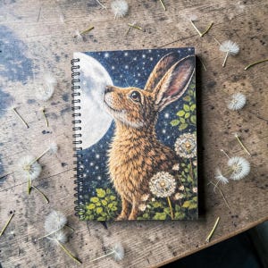 May include: Spiral-bound notebook featuring a detailed illustration of a brown hare gazing at a full moon against a starry night sky. Dandelions and foliage frame the hare. The notebook rests on a rustic wooden surface scattered with dandelion seed heads.
